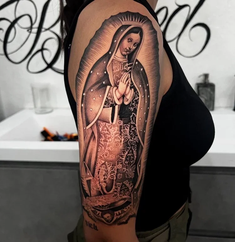 Tattoo of the Virgin of Guadalupe on a person's upper arm, depicting a religious figure with hands clasped in prayer, surrounded by a halo, with detailed robes and a background of stars.