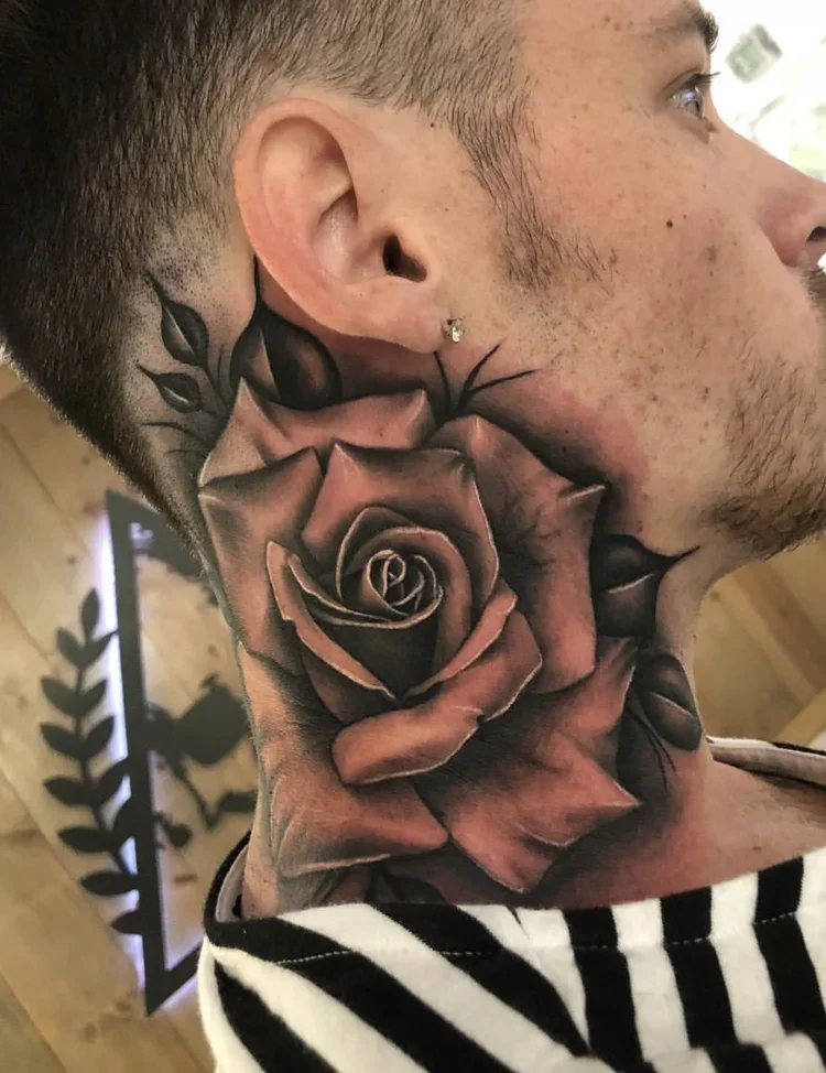 Close-up of a man's face and neck tattoo featuring a large, detailed rose with shaded petals, behind the ear.