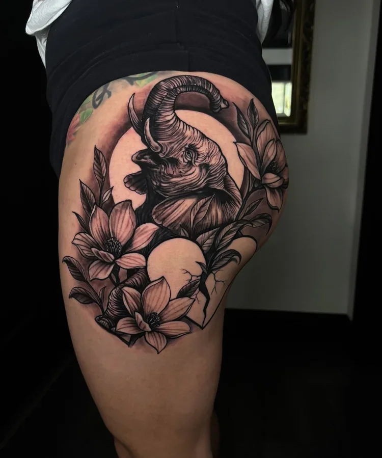 Tattoo of an elephant with curved trunk, surrounded by flowers and leaves, with a moon and starry background on a person's thigh.