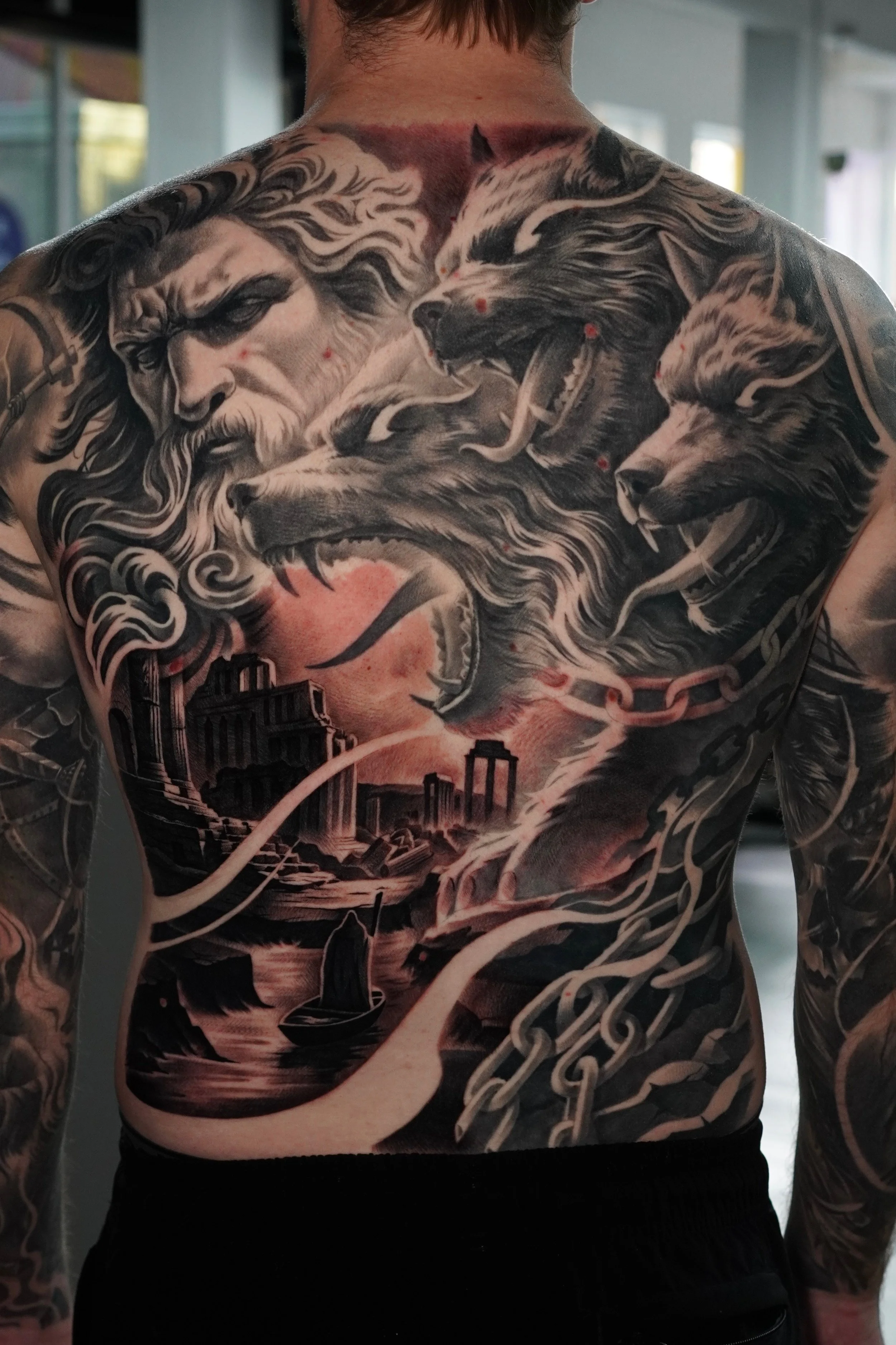 A person's back and chest covered in detailed black and gray tattoos, including a fierce wolf's head, a lion's face, a woman with long flowing hair, cityscape, clouds, and chains.