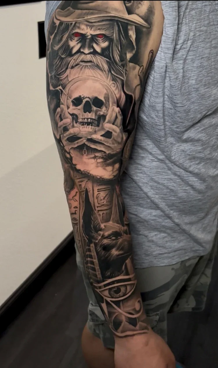 Black and gray tattoo of a bearded man with a hat holding a skull, with an Egyptian eye symbol, hieroglyphics, and a dog's face running down the forearm.
