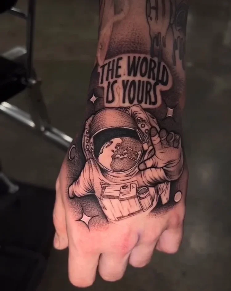Tattoo on a person's hand features a hand holding the Earth, an astronaut helmet, stars, and the phrase 'The world is yours'.