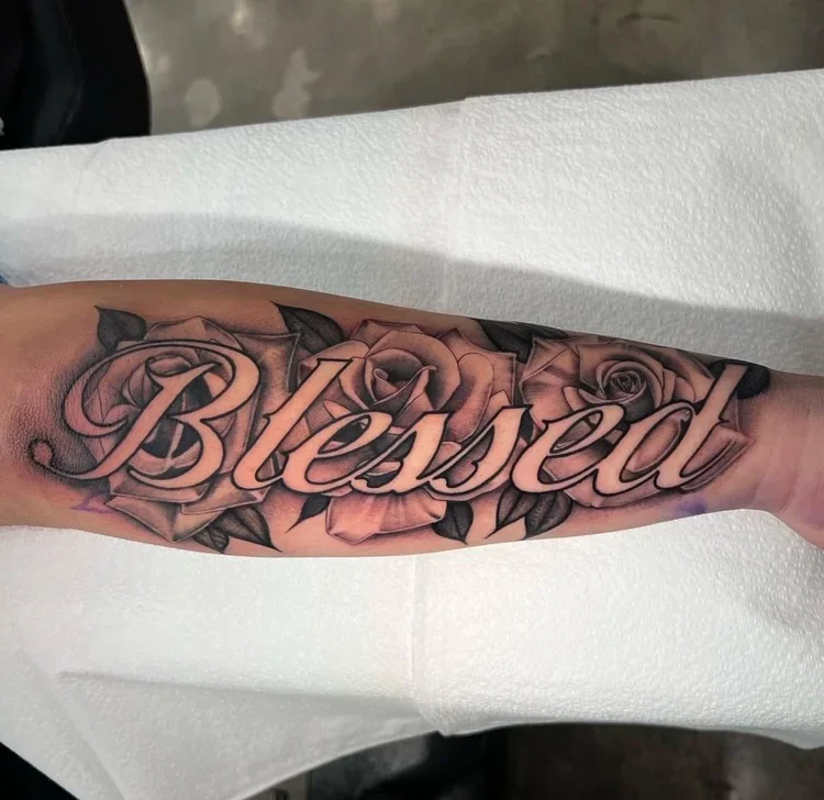 Tattoo of the word "Blessed" in cursive script with roses in the background on a person's forearm.