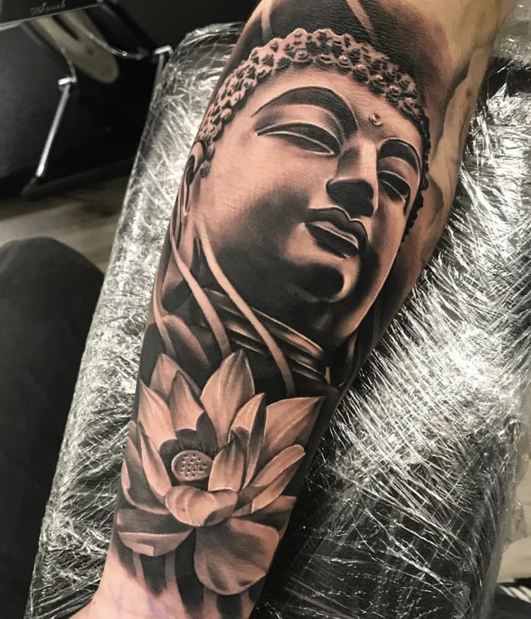 Black and gray tattoo of a serene face of Buddha with a lotus flower at the base on a person's arm.