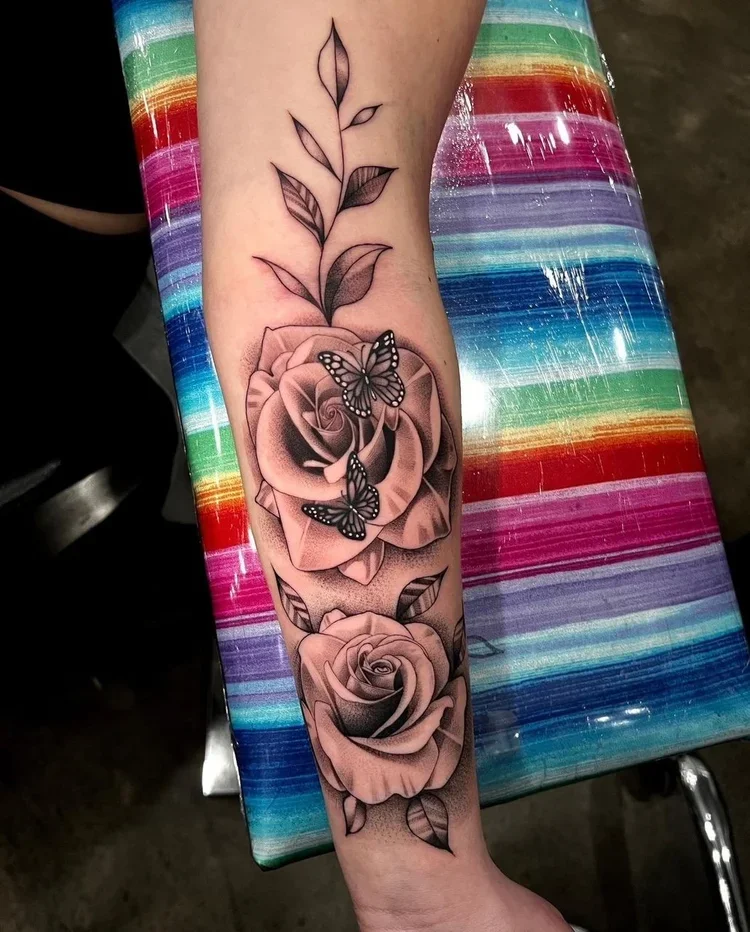 Tattoo of two roses with butterflies on a person's forearm, with leaves extending upward. The tattoo is detailed in black and gray shading, set against a rainbow-striped surface.