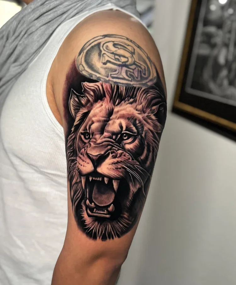 Tattoo of a lion's head with a fierce expression on a person's upper arm, with a coin over the shoulder.