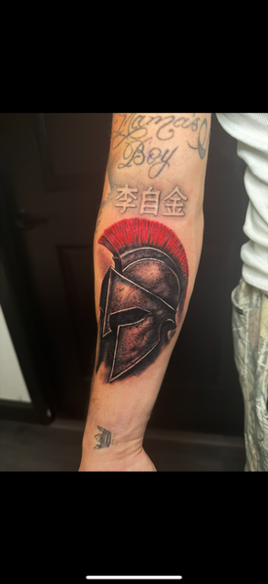Tattoo of a Spartan helmet with a red crest on the forearm, with Chinese characters below and script above.