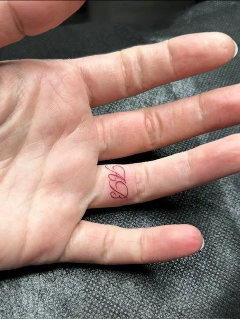 A close-up of a person's left hand resting on a dark textured surface, showing a red ink tattoo of a small, cursive number 3 on the ring finger.