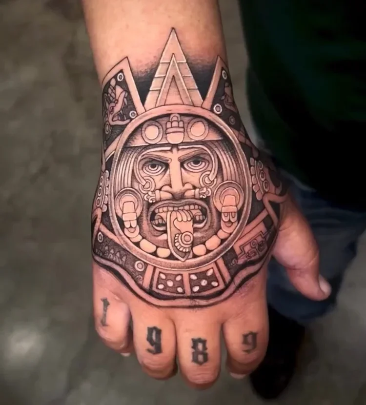Tattoo of an Aztec warrior face with intricate designs on the hand and fingers, with the year 1999 tattooed on the fingers.