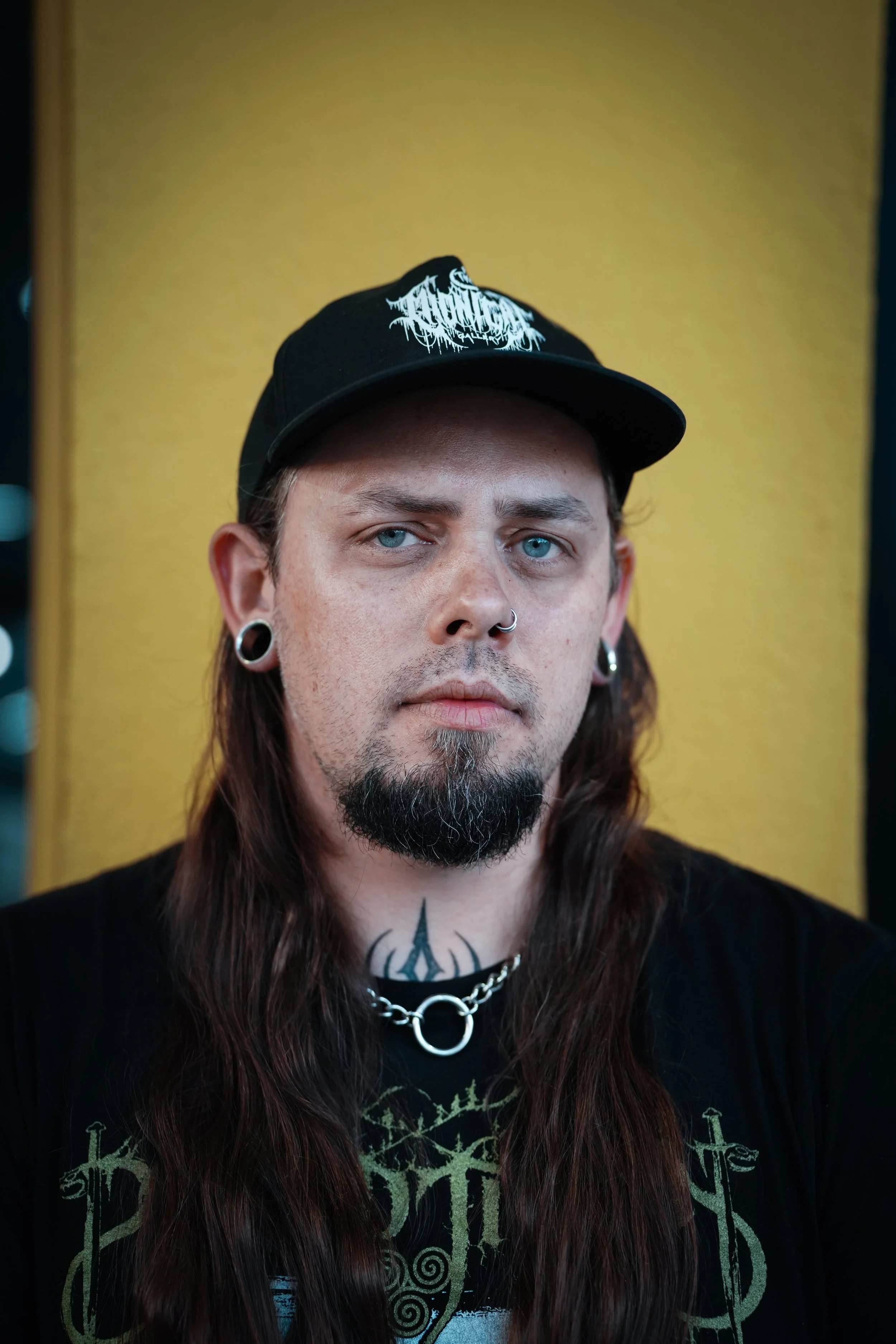 A man with long brown hair, wearing a black cap with white text, black shirt with green graphics, silver chain necklace with ring, and earrings, standing in front of a yellow background.