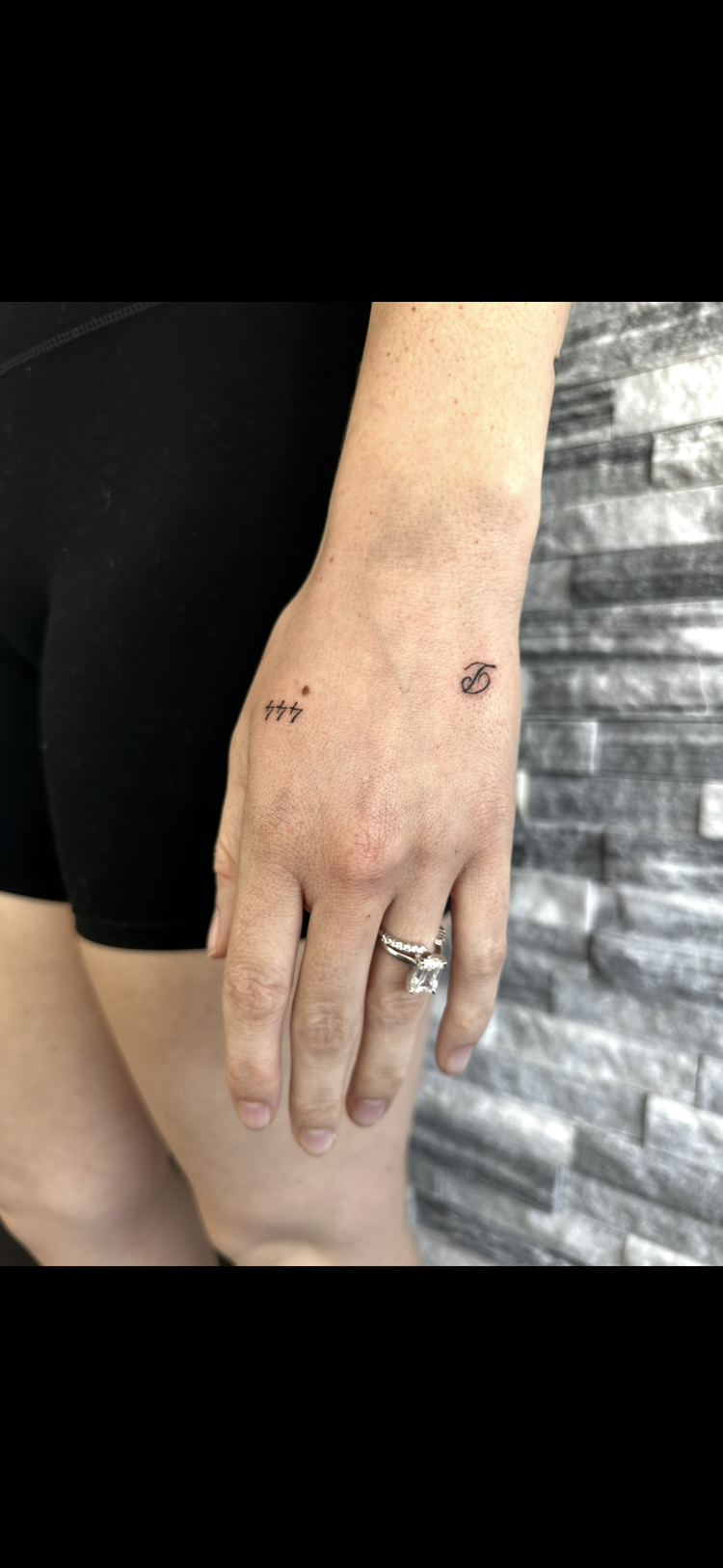 A person's left hand resting on their thigh, showing small tattoos on the wrist and finger, with the person wearing a silver engagement ring with a large gemstone.