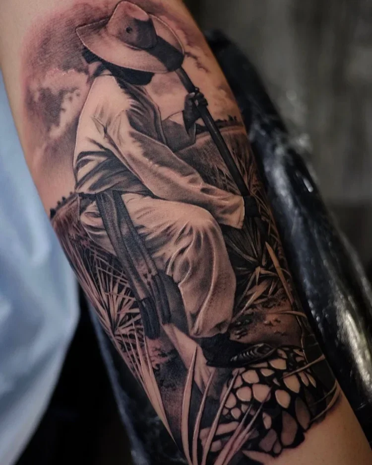 Black and gray tattoo of a person sitting on a chair, wearing a wide-brim hat, face turned downward, with detailed surrounding foliage and grass.