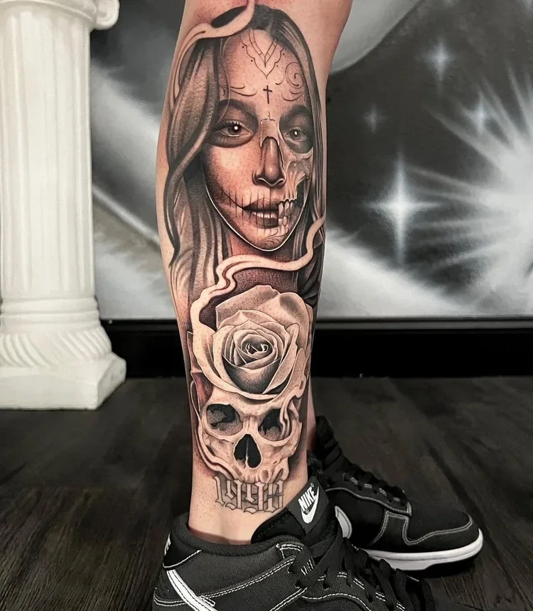 Tattoo on lower leg featuring a woman's face with half of the face as a skull, a rose, and decorative elements.