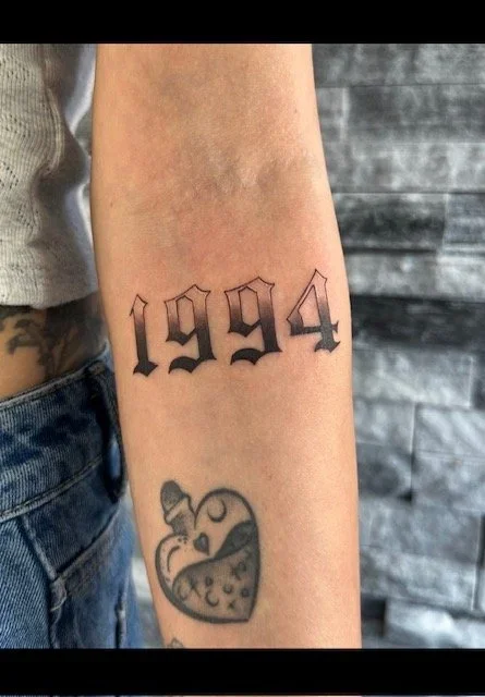 Tattoo on person's forearm displaying the year 1994 in black gothic font above a small, colorful heart with a skull and bunny design inside it.