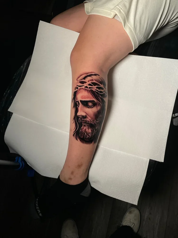 Tattoo of Jesus Christ with a crown of thorns on a person's calf.
