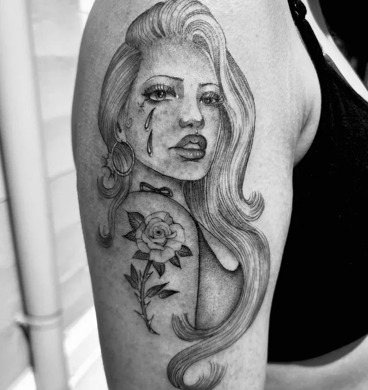 Black and white tattoo of a woman with long hair, tears on her face, and a rose on her shoulder.