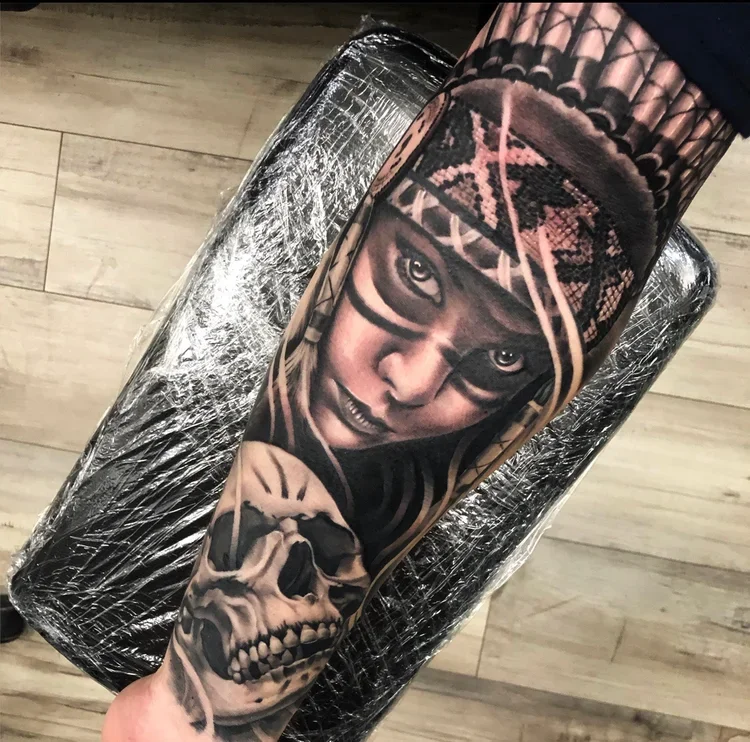 A detailed black and gray tattoo of a woman in tribal headdress with piercing eyes, holding a skull, on a person's arm.