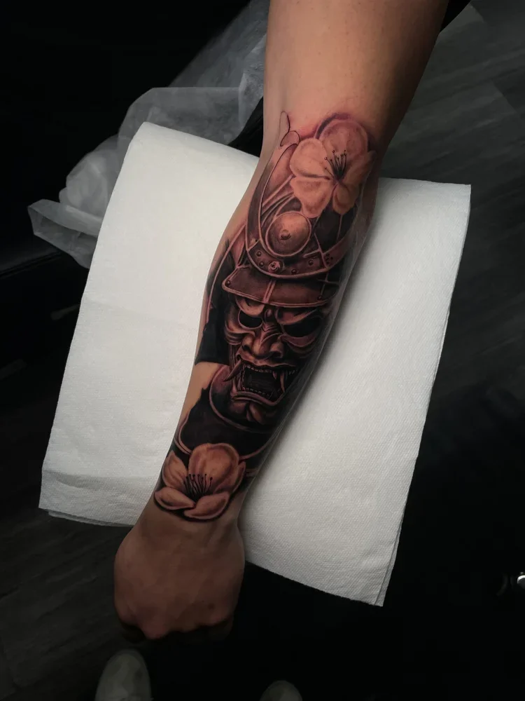 Tattoo of a fierce samurai mask with cherry blossoms on a person's forearm.