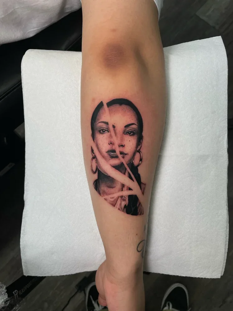 A black and gray portrait tattoo of a woman with hoop earrings, short hair, and distinctive facial features. The tattoo has an artistic, stylized appearance, with some elements like the woman’s finger and hair blending into abstract shapes.