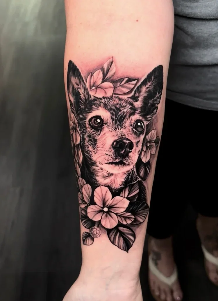 Black and gray tattoo of a dog surrounded by flowers on a person's forearm.