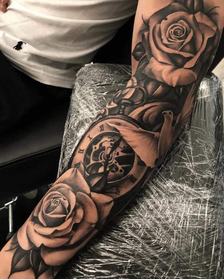 Detailed black and gray tattoo on a person's forearm featuring a rose, pocket watch, and a bird in flight.