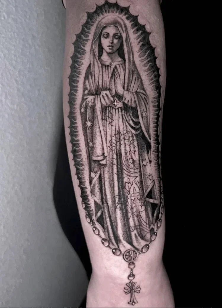Black and gray tattoo of the Virgin Mary with hands in prayer, surrounded by a religious rosary with a cross at the bottom