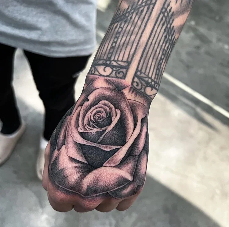 Close-up of a tattooed hand featuring a detailed black and gray rose on the back of the hand, with part of a decorative fence or gate design visible above.