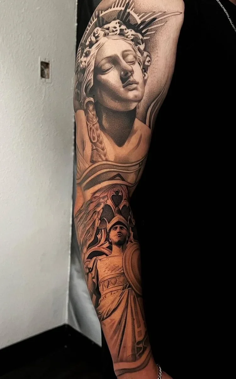 Black and gray tattoo of a woman's face with closed eyes, a crown, and flowing hair on the upper arm, and a warrior woman with a shield and armor on the lower arm.
