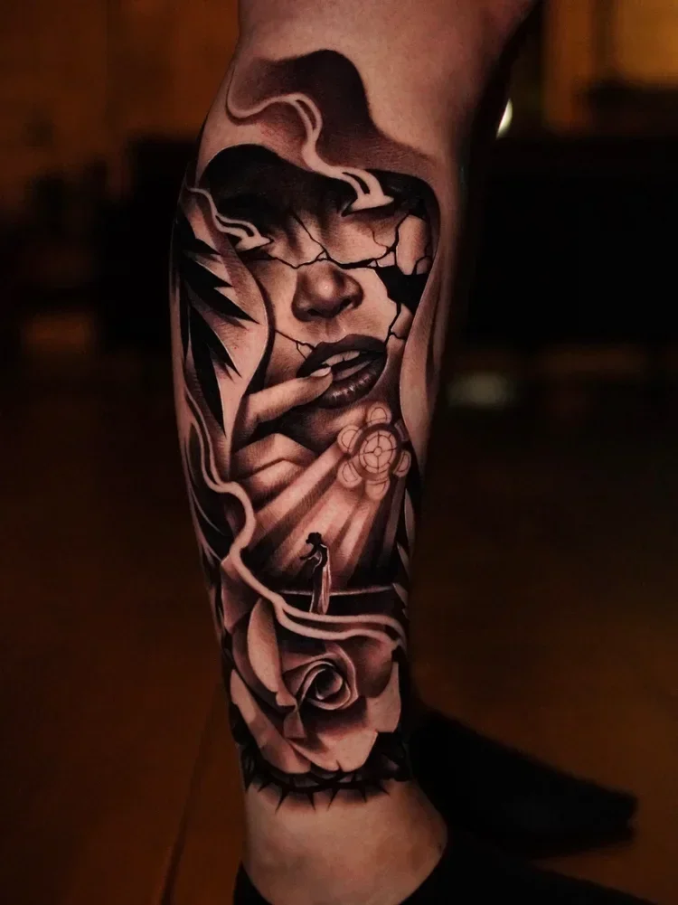 Tattoo of a woman's face with lips slightly parted, cracking as if shattering, with hands near her face, a rose, and abstract patterns in black and gray on a person's arm.