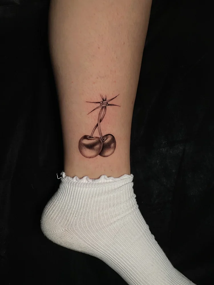 Tattoo of cherries tied together with a barbed wire design on a person's leg.