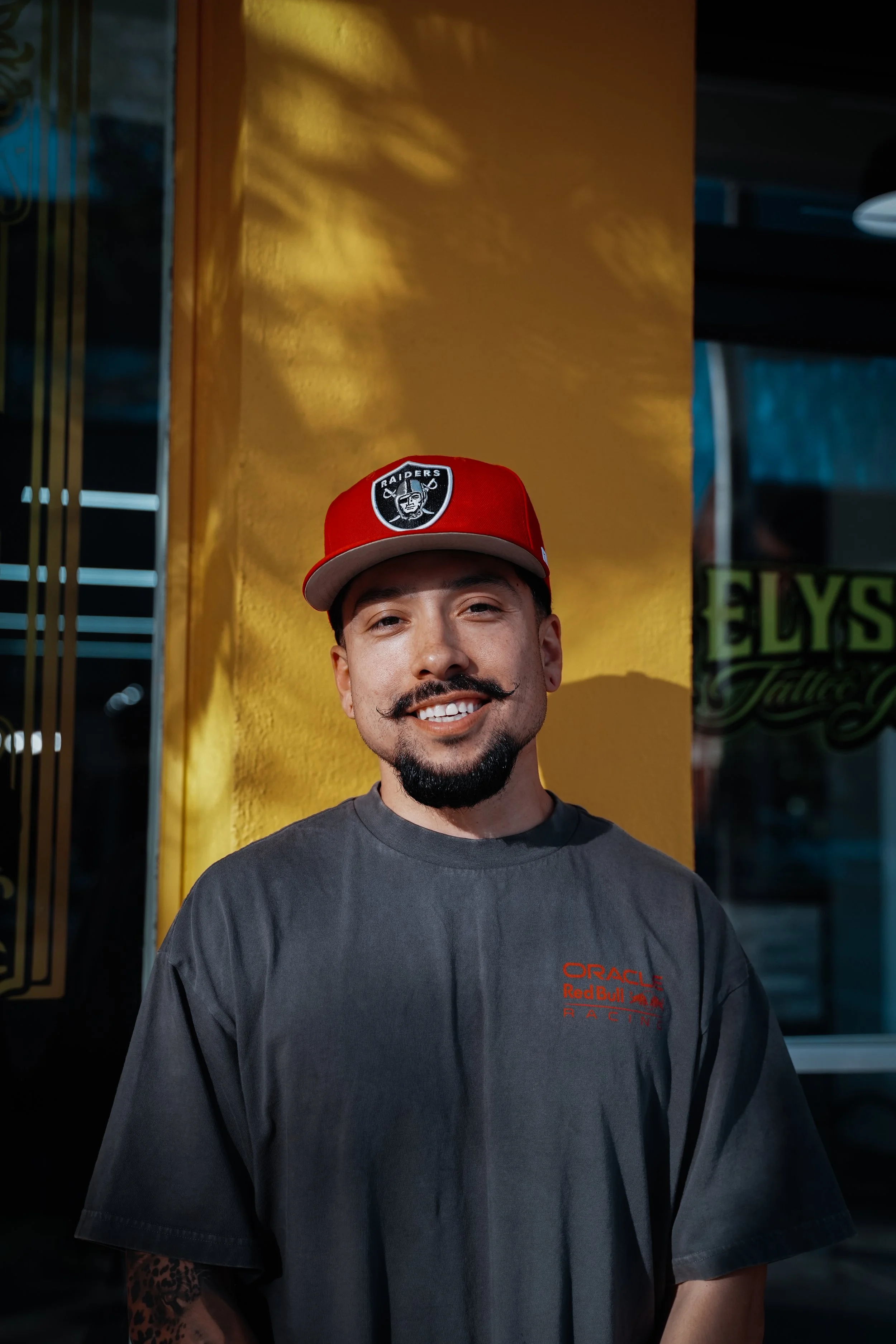 A man with a goatee and mustache smiling, wearing a red Raiders cap and a dark gray Oracle Red Bull Racing T-shirt, standing in front of a yellow wall with a glass window reflecting the street.