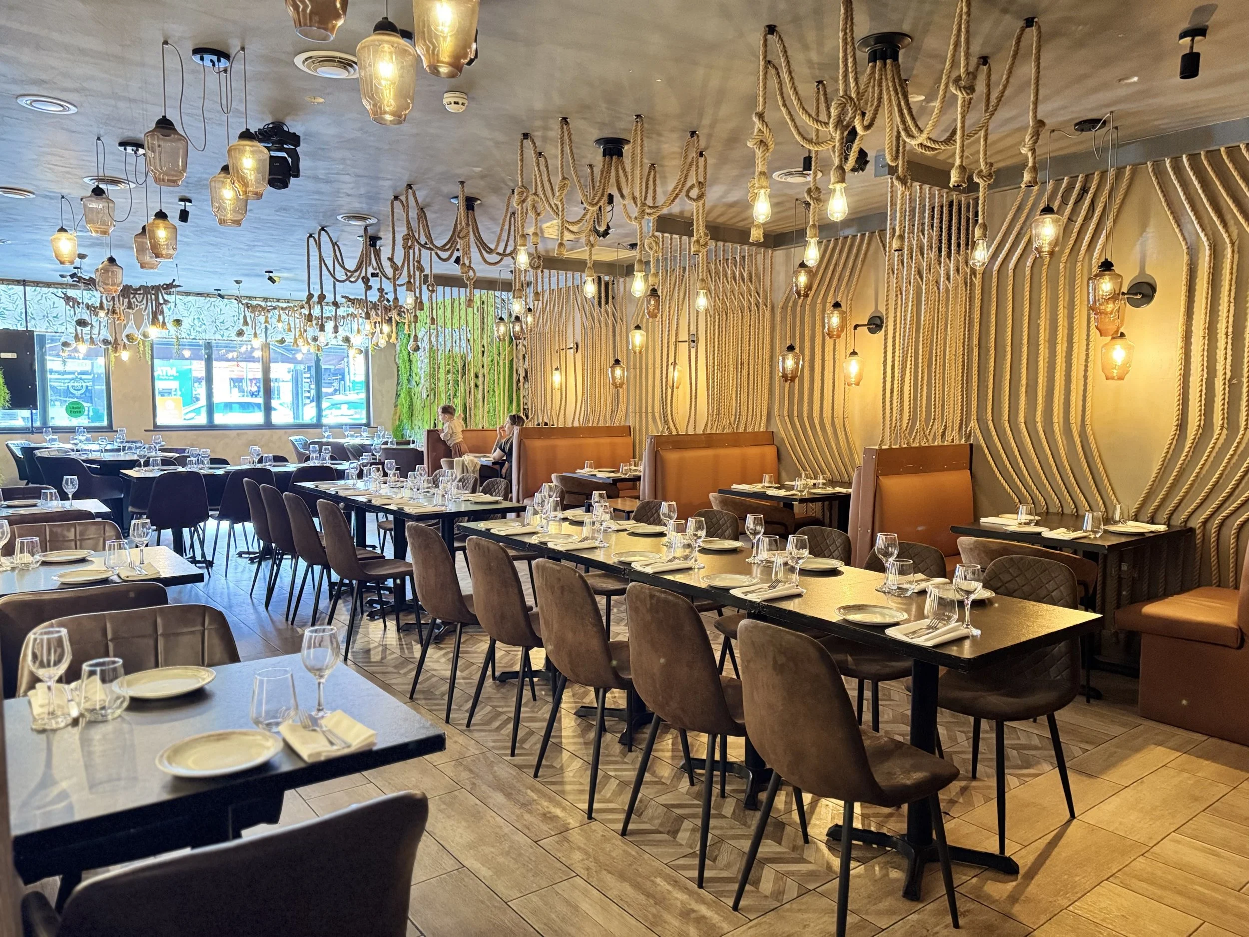 Interior of a modern restaurant with wooden flooring, set tables with plates, glasses, and napkins, and decorative hanging lights and ropes on the walls.