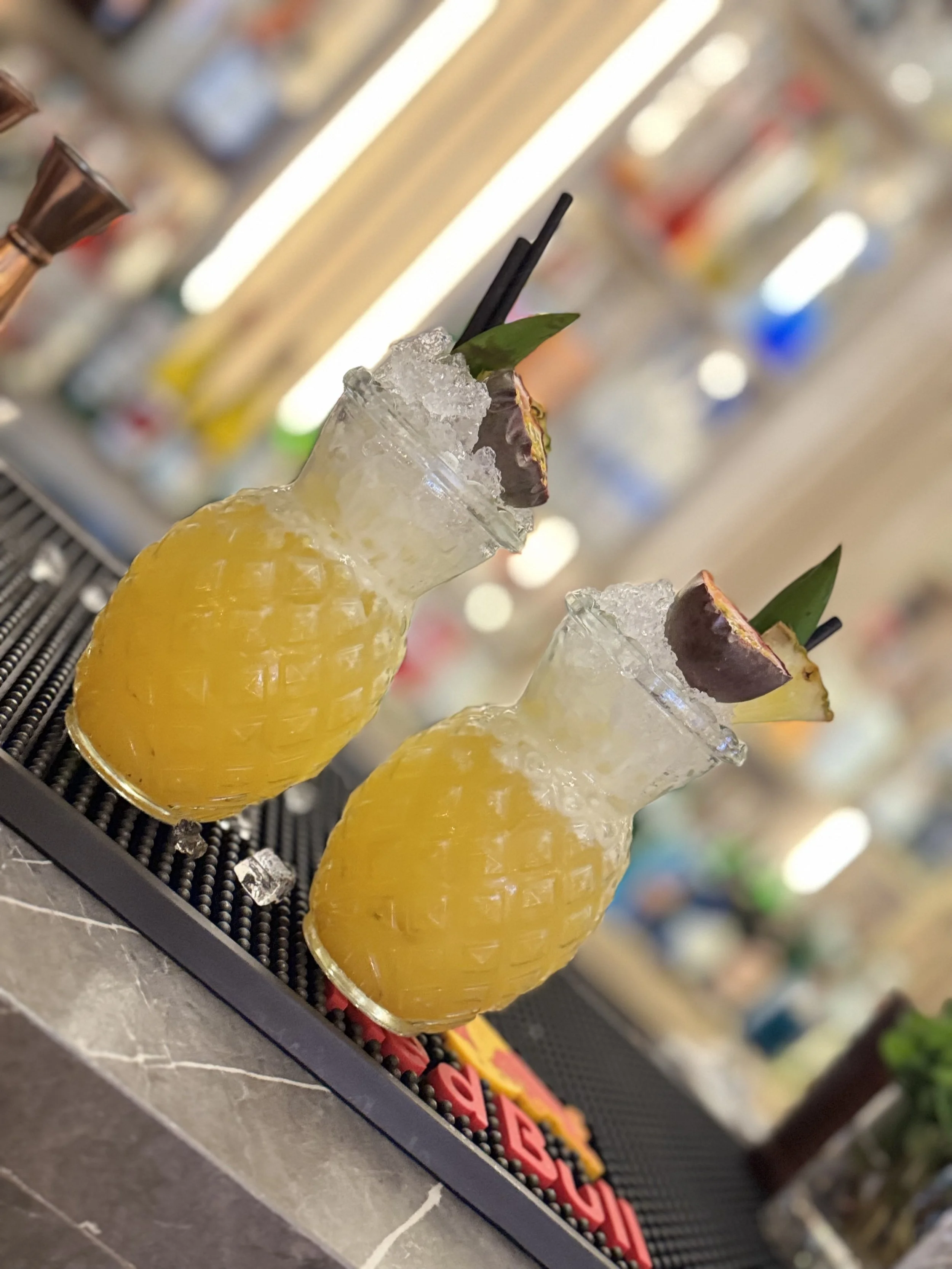 Two pineapple-shaped glasses filled with a yellow cocktail, garnished with pineapple slices, lime leaves, and cherries, and topped with crushed ice.