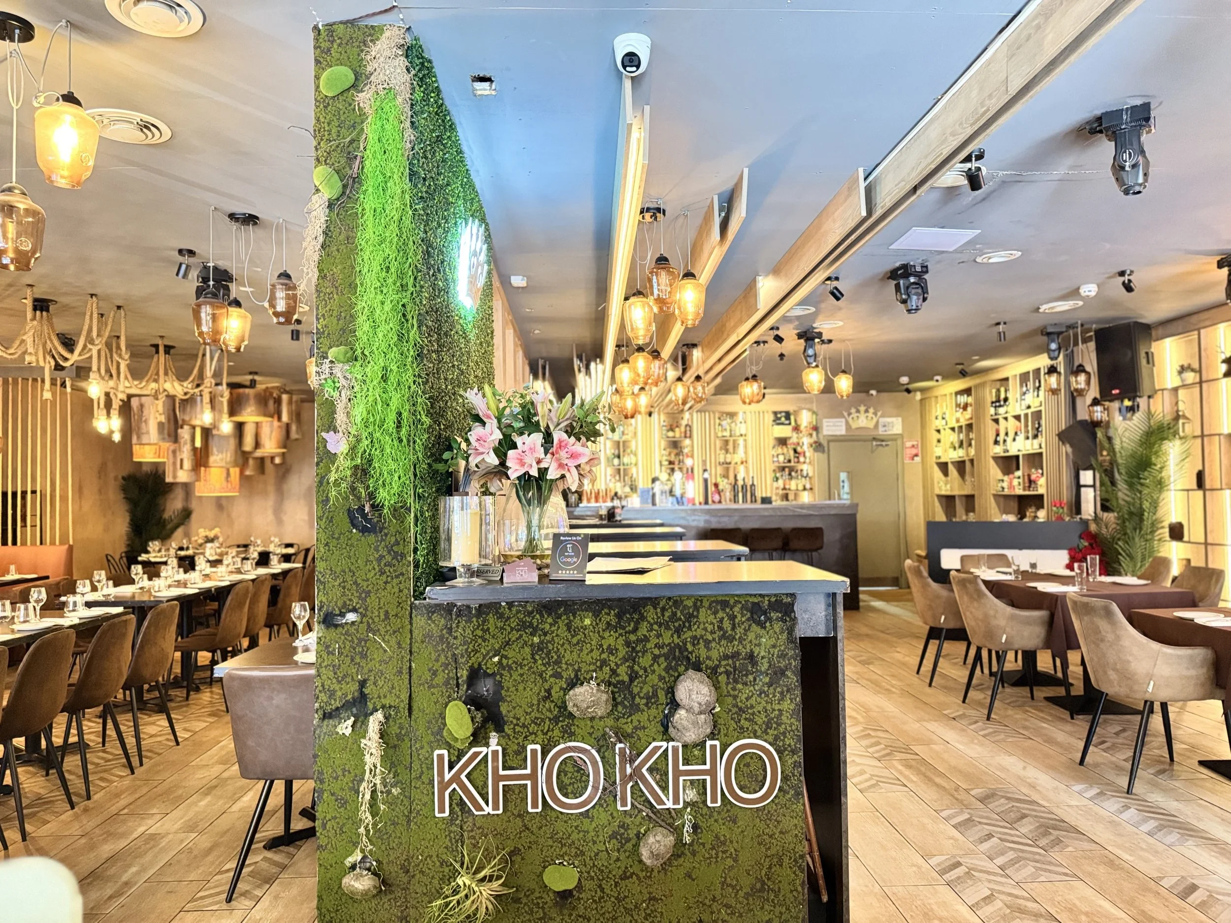 Interior of a restaurant with dining tables, chairs, green wall decor, floral arrangements, warm lighting, and a bar area in the background.