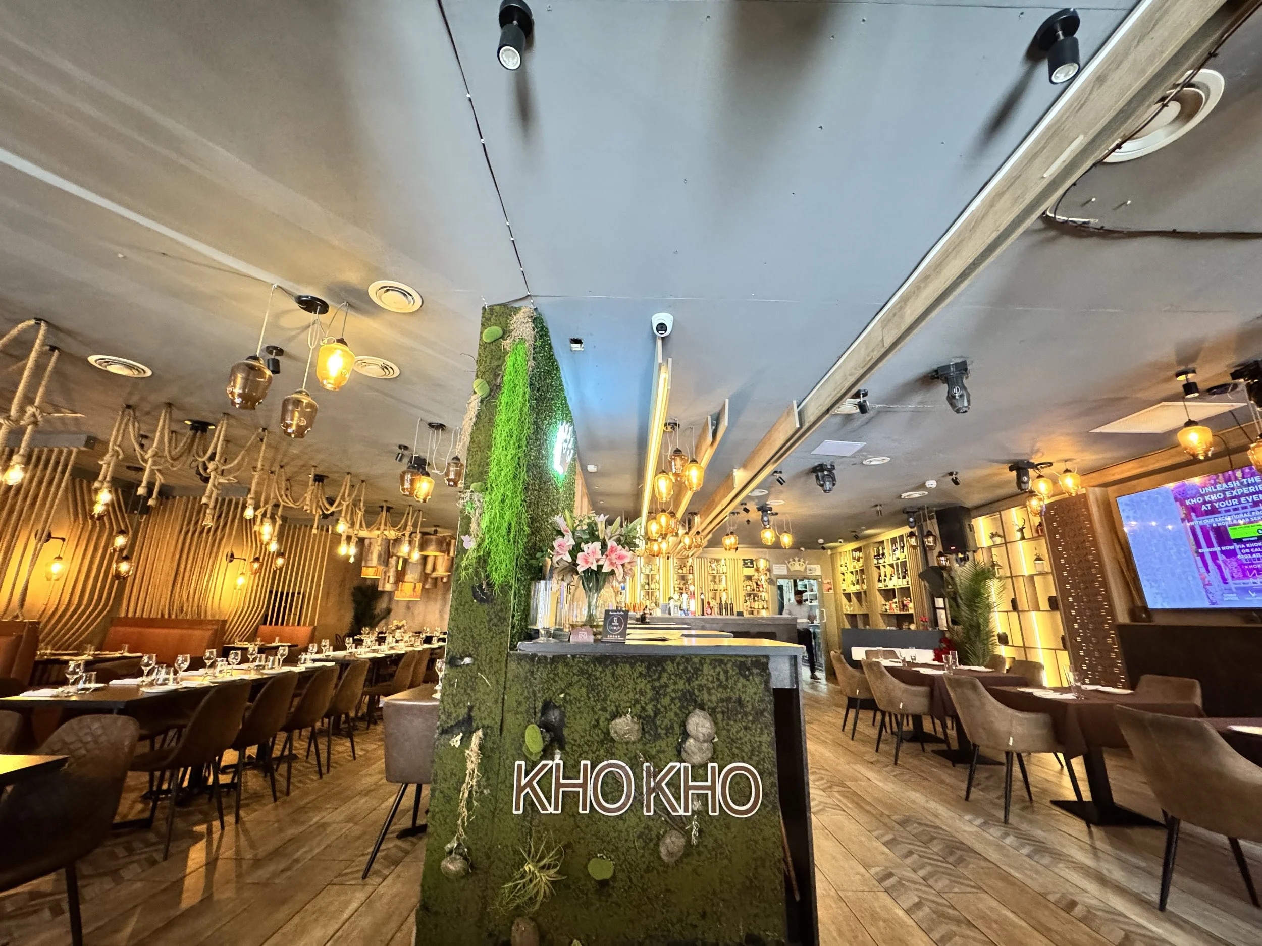 Interior view of a modern restaurant or cafe with wooden flooring, stylish chairs, and warm lighting. A moss-covered partition with a sign reading 'KHO KHO' separates the space. The background features shelves with decor, a large digital screen, and 