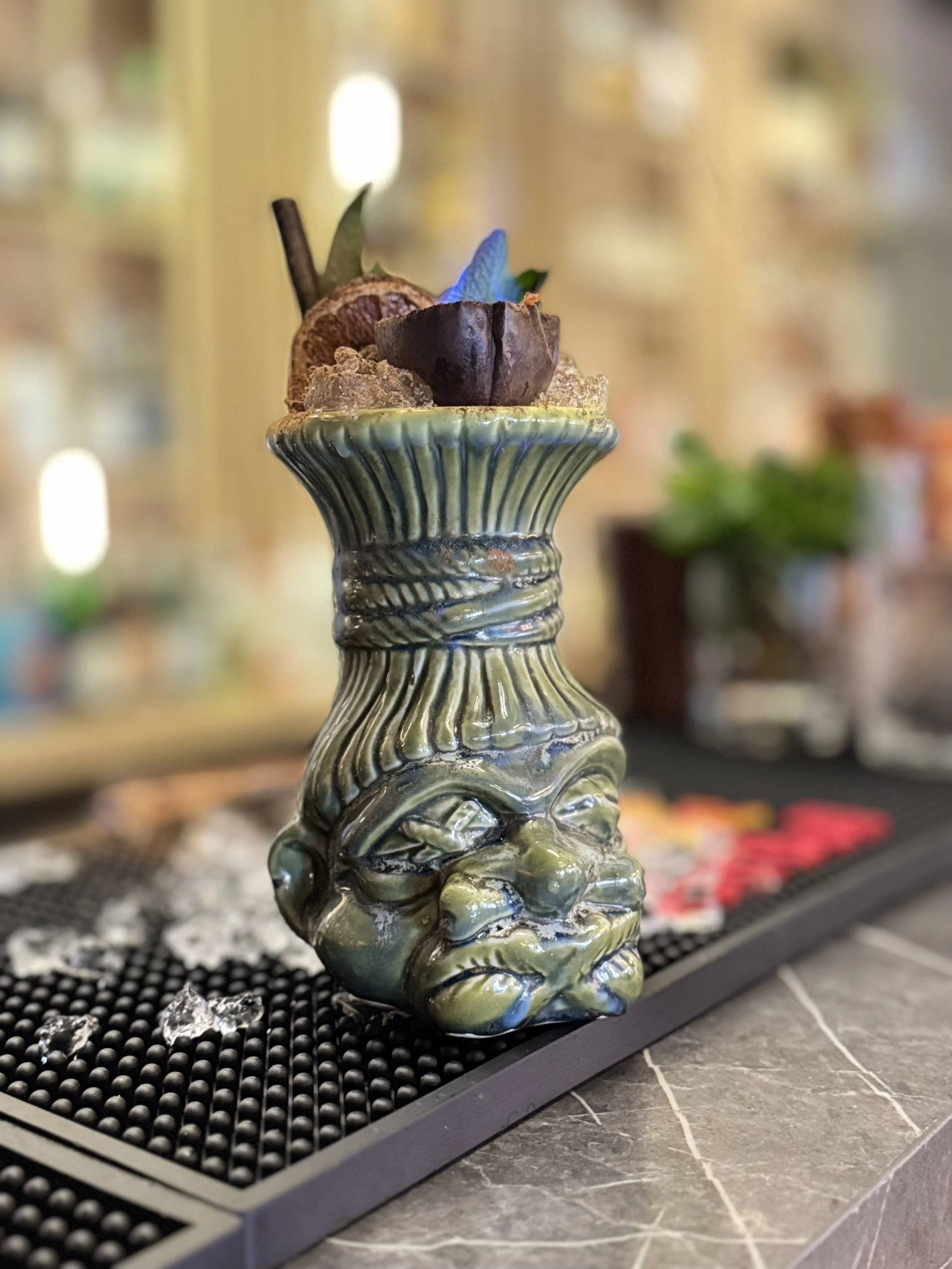 A ceramic vase with a face decoration, holding a plant, placed on a bar counter with scattered ice and a bar mat.