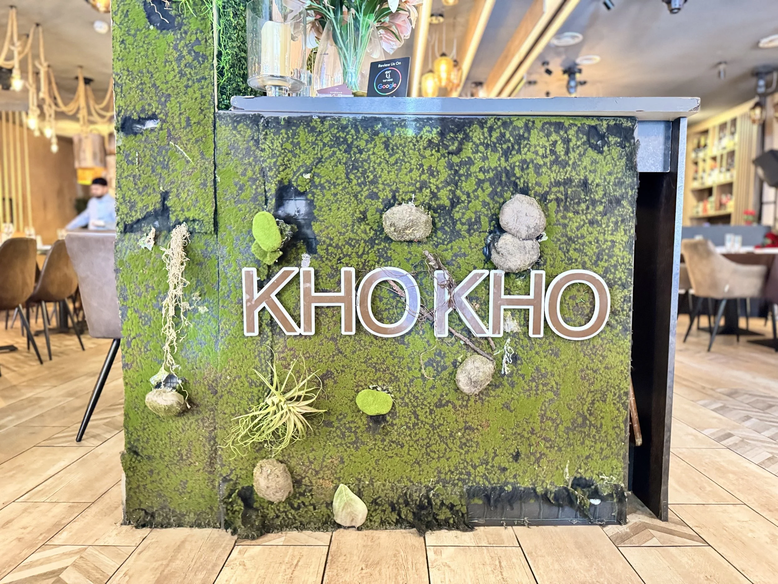 Close-up of a moss-covered, green and black textured wall with various small, round, and irregularly shaped objects attached, and the text 'KHOKHO' in bold white letters.