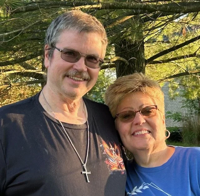 A man and woman standing close together outdoors, smiling, with green trees and sunlight in the background.