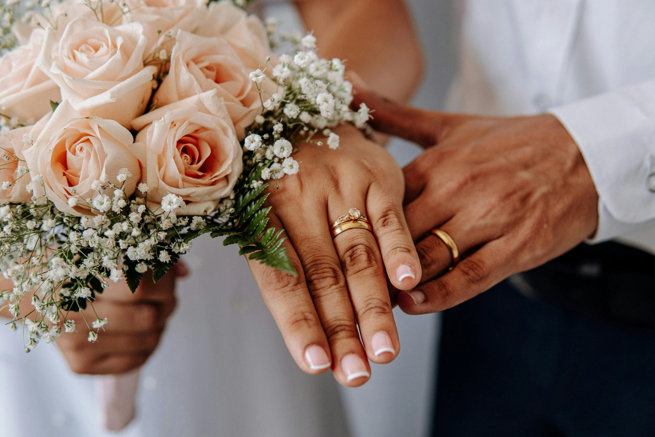 Close-up of two hands, one with a gold ring, clasped over a bouquet of light peach roses and white baby's breath flowers.