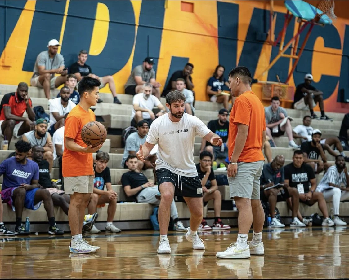 A basketball coach demonstrates a move to two players during a practice session on an indoor court with spectators in the background.
