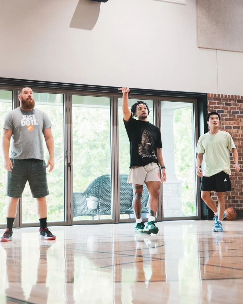 Three men in a gym, with one holding a basketball overhead, casually dressed, standing on a polished wooden floor with large windows behind them.
