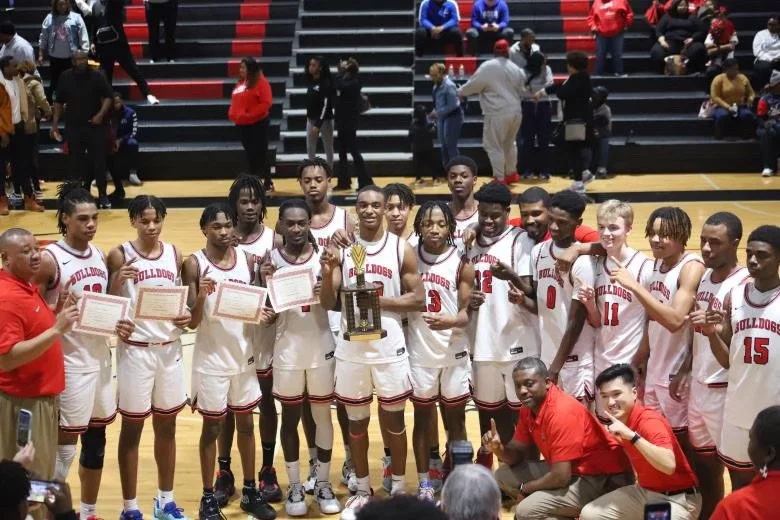 Basketball team in white jerseys with red accents celebrating on court, holding awards and a trophy, with spectators in the background.