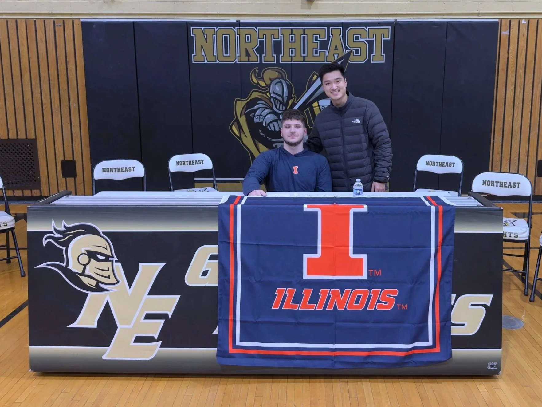 Two young men in a gymnasium at a signing event with an Illinois basketball player, seated at a table with an Illinois flag and signed paper, with empty chairs labeled 'NORTHEAST' behind them and a logo on the wall.