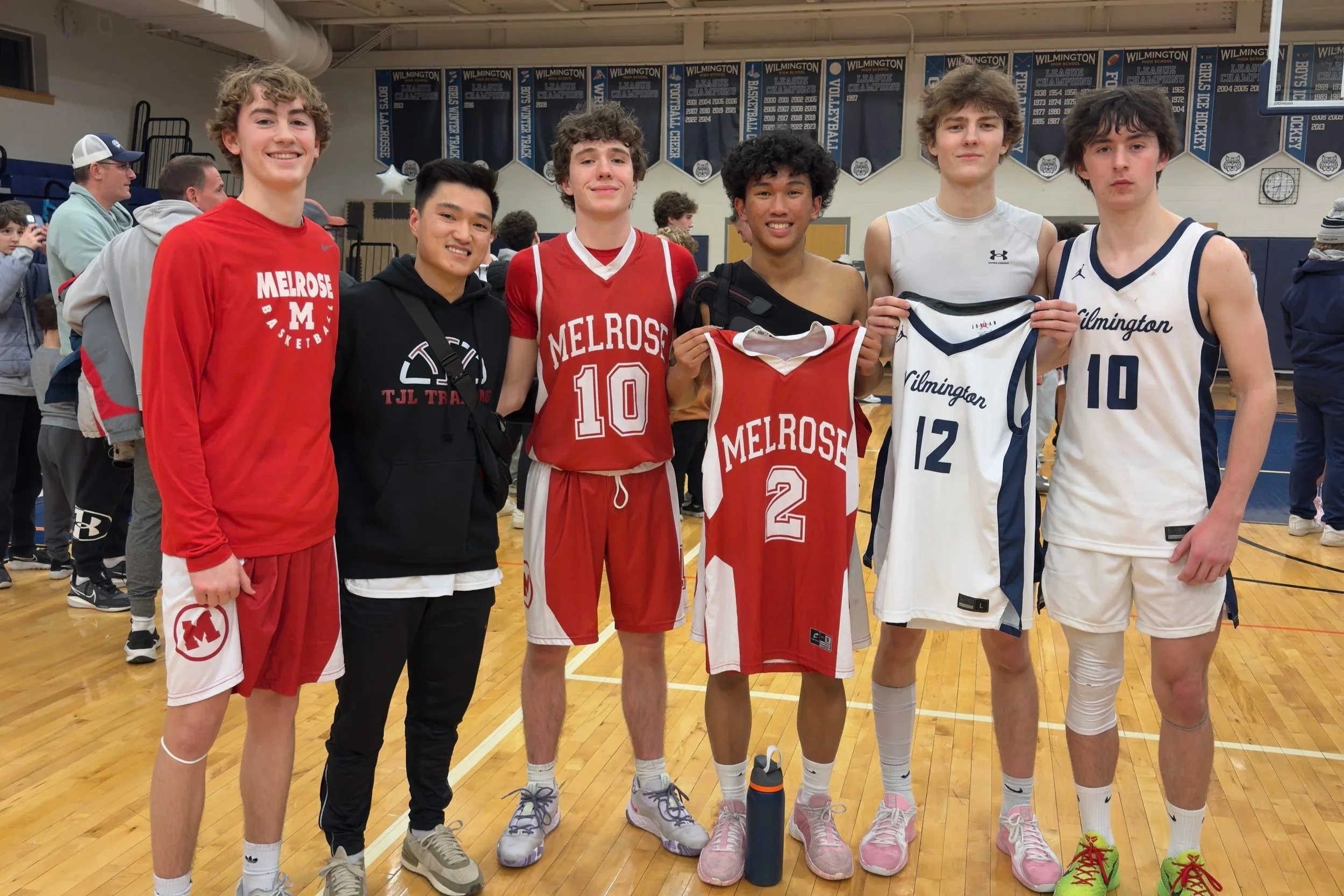 Group of six boys in a gymnasium, wearing basketball uniforms and casual clothes, holding basketball jerseys from Melrose and Wilmington teams, smiling at the camera.