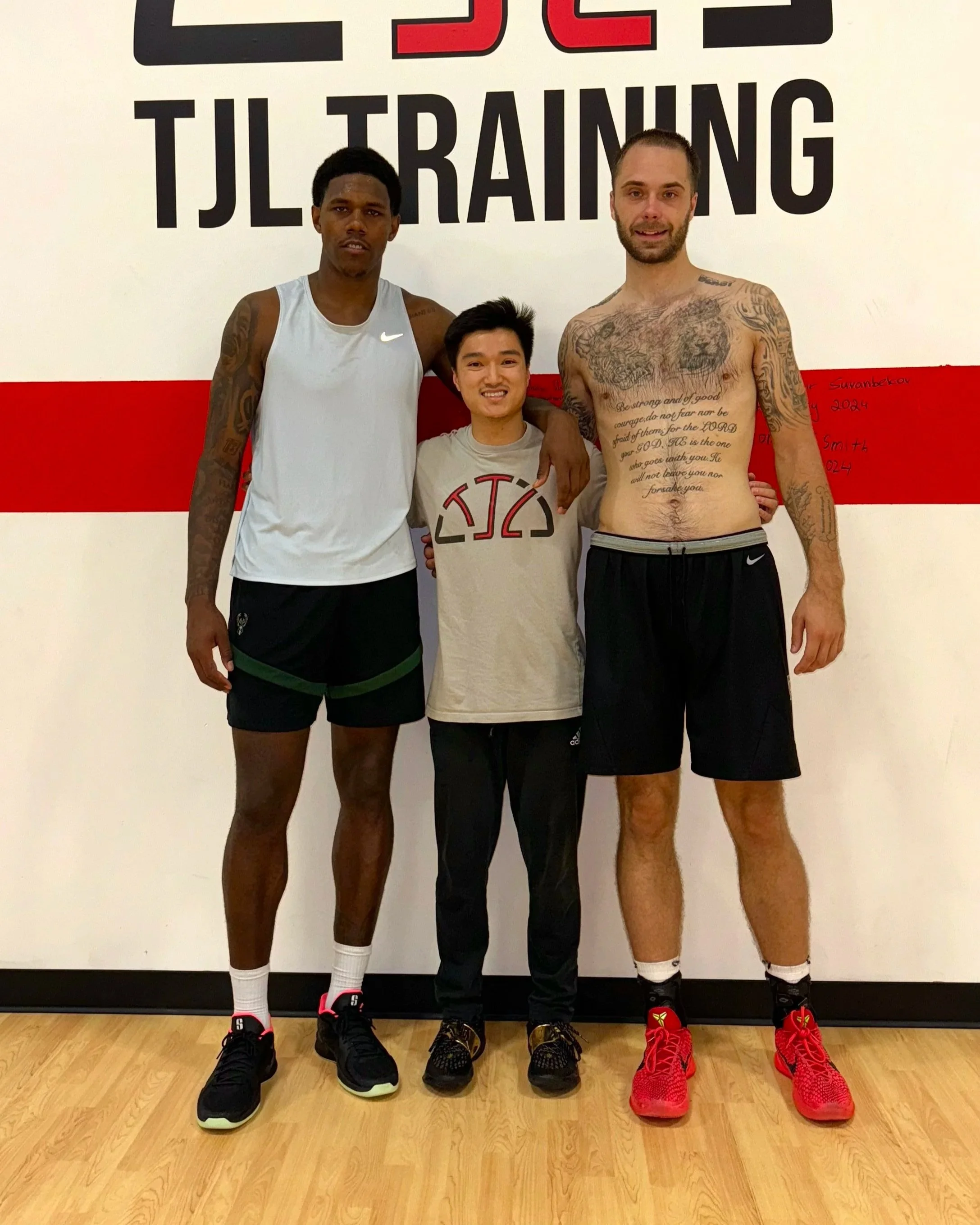 Three men standing side by side in workout clothing in front of a wall with training-themed text. Two tall men are on either side, and a shorter man is in the middle.