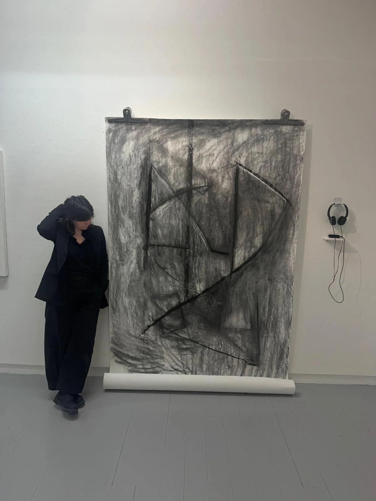 A woman in black clothing stands next to an abstract black and white charcoal drawing on paper, mounted on a wall with clips, in an art gallery. A pair of headphones hangs on the wall beside the artwork.