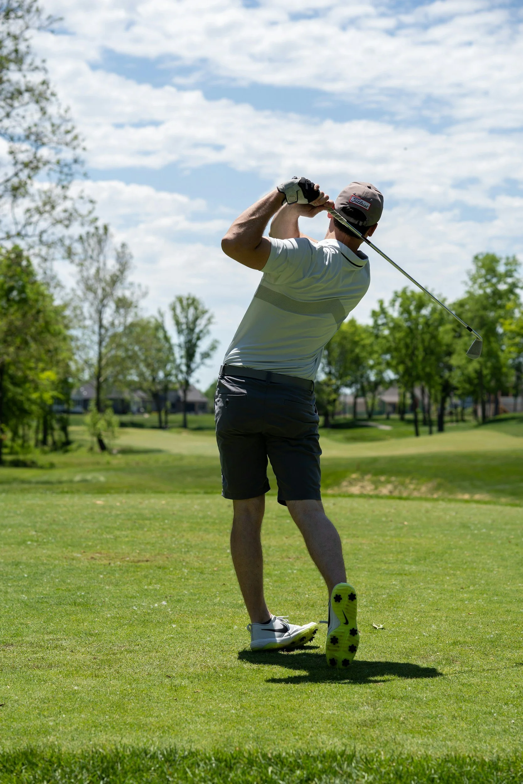 A man in a golf course swinging a golf club, wearing a gray cap, white shirt, dark shorts, and athletic shoes, with green trees and a blue sky with some clouds in the background.