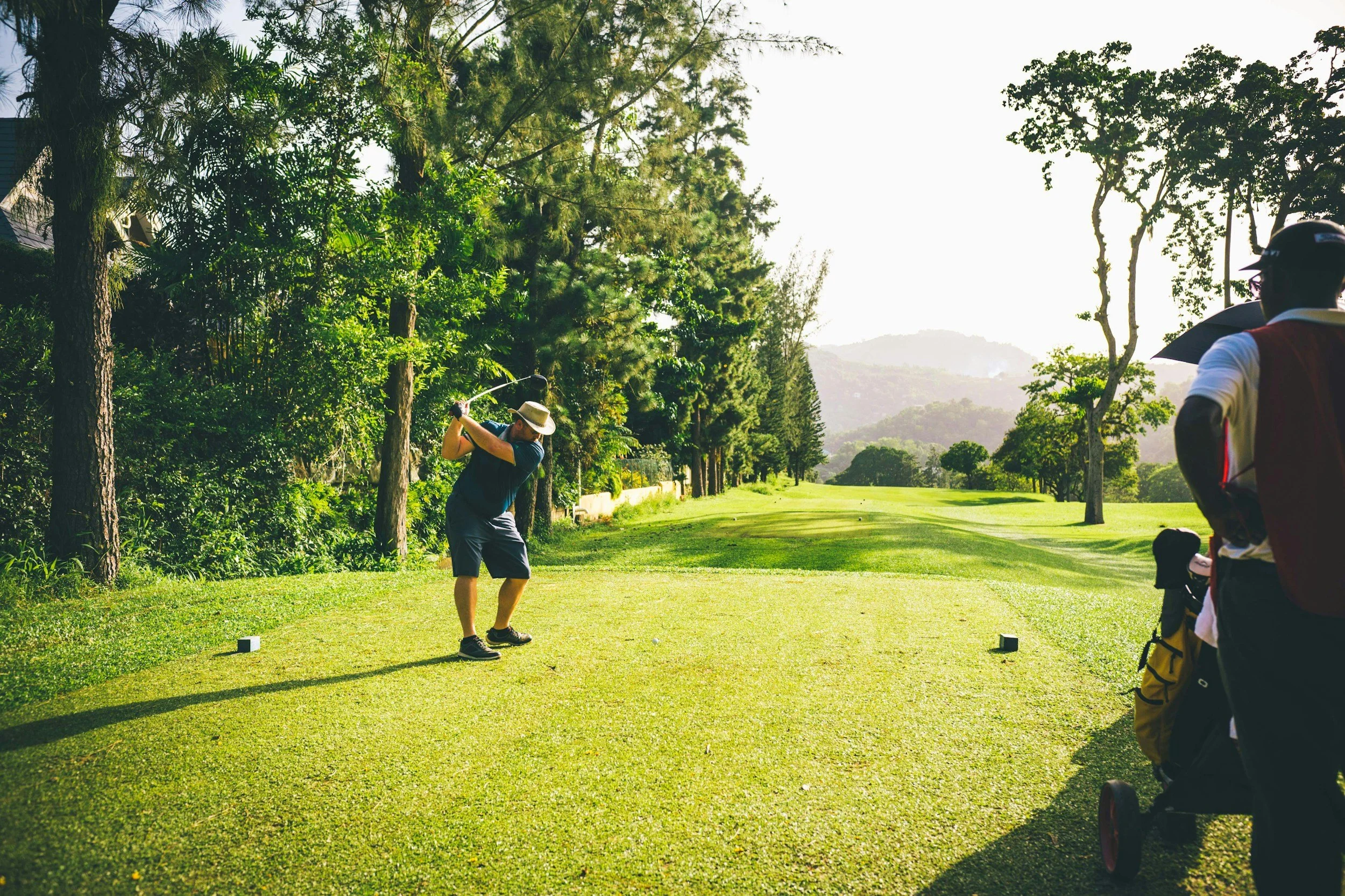 Two men playing golf on a lush green golf course surrounded by trees, with a mountain in the background and a person holding an umbrella on the right.