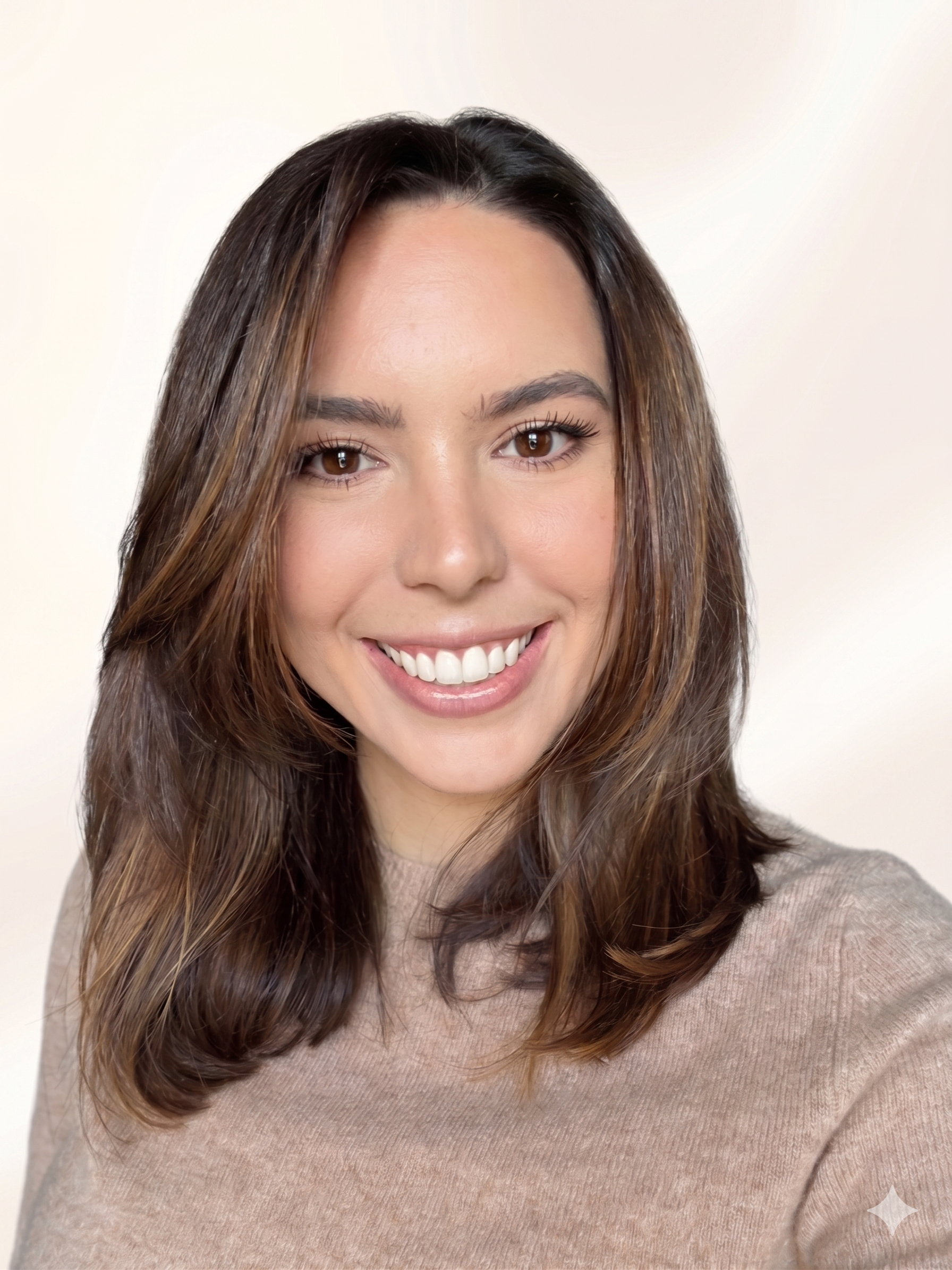 Close-up of a woman with long, dark brown hair, smiling, wearing a beige sweater, against a plain light background.