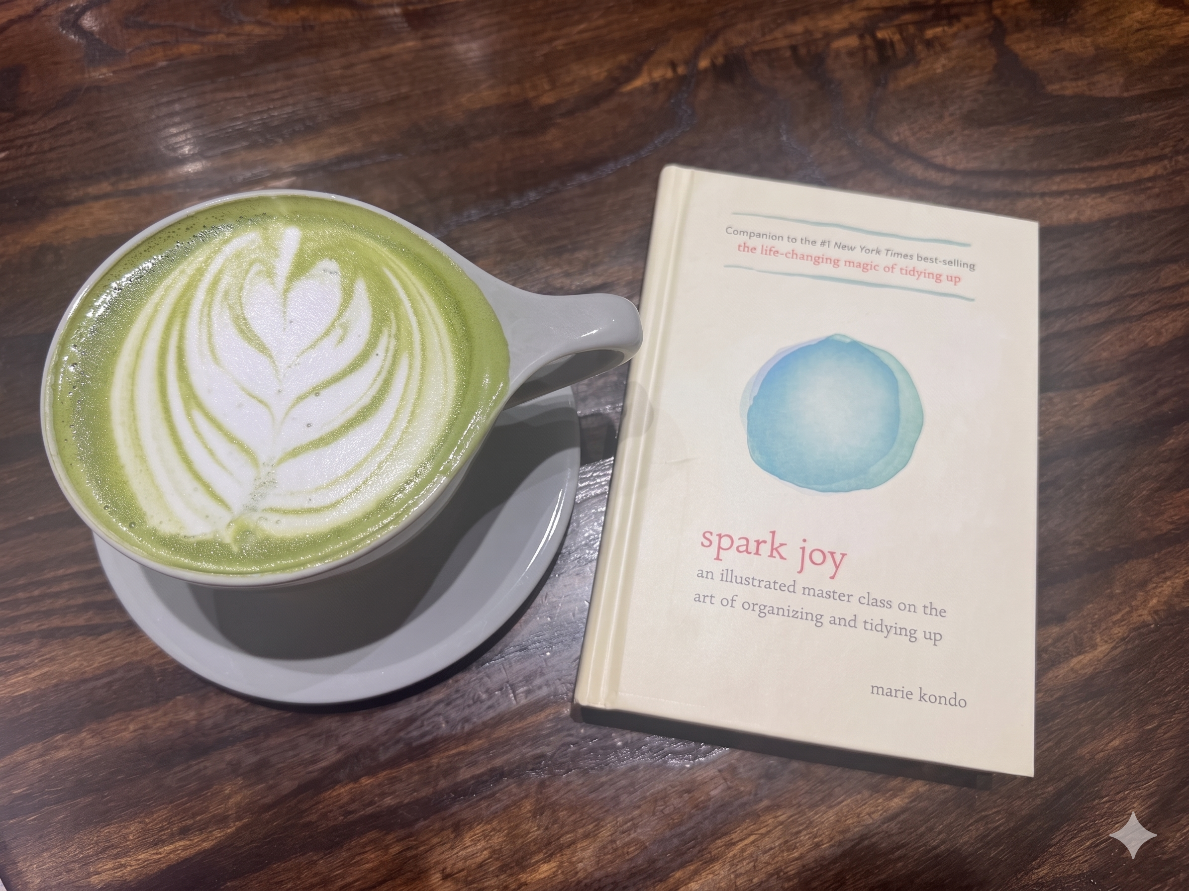 A cup of matcha latte with latte art in the shape of a heart, placed on a wooden table next to a book with a watercolor illustration of a blue circle on the cover.